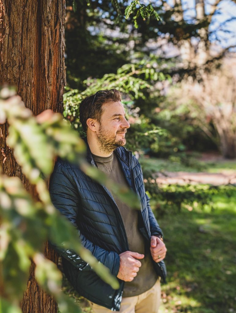 A man leans against a tree, smiling, holding his coat with both hands in Villa dei Cedri Park in Bellinzona