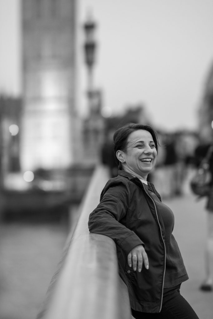 A woman leans against the railing of a bridge in London, in front of Big Ben, laughing